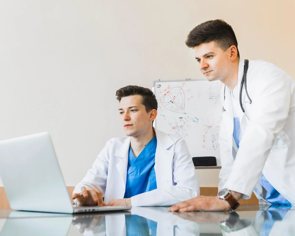 Two medical professionals in white coats collaborate over a laptop, with a whiteboard filled with diagrams in the background