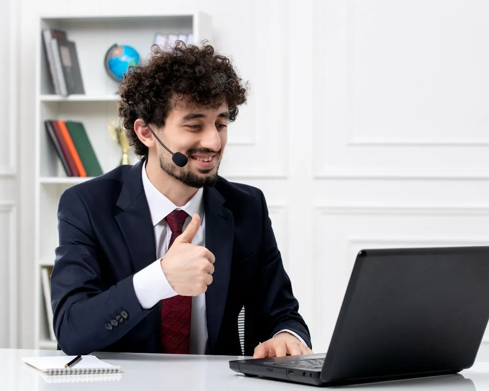 A man in a suit gives a thumbs-up while sitting at a desk with a laptop